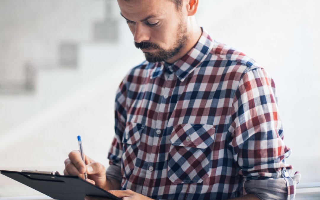 A man in a plaid shirt writes notes on a clipboard, focused on his task